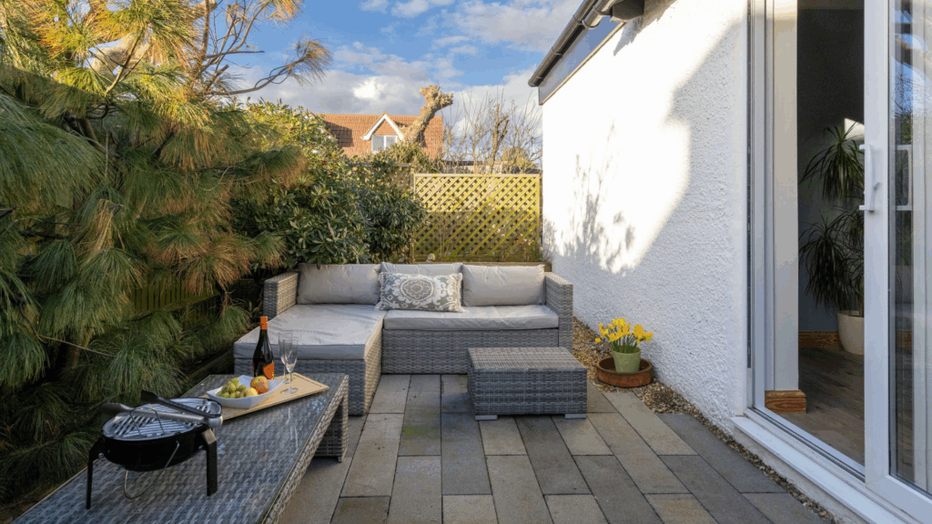 A sunny patio area with a grey rattan outdoor sofa. On the table is a small round barbecue, a bottle of wine with an orange label, two wine glasses and a fruit bowl.