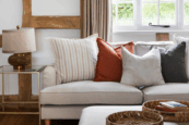 A cozy living room corner with a beige sofa decorated with earthy-toned cushions, a glass-topped side table with a gold lamp, wooden beams, neutral curtains, and a woven tray with books on an ottoman.