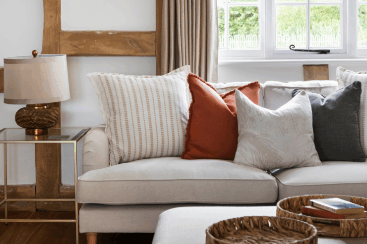 A cozy living room corner with a beige sofa decorated with earthy-toned cushions, a glass-topped side table with a gold lamp, wooden beams, neutral curtains, and a woven tray with books on an ottoman.