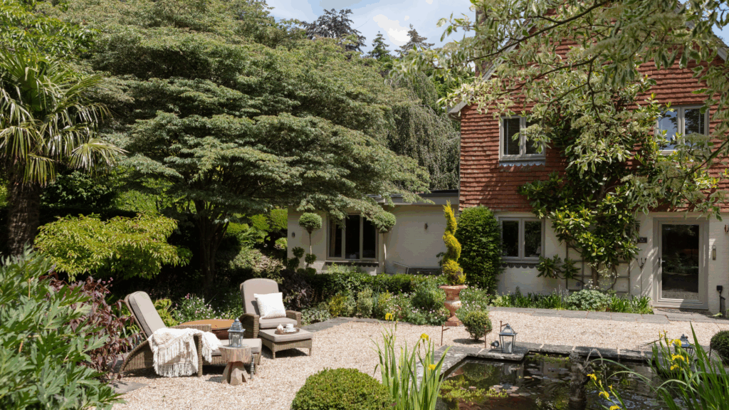 A charming garden with lounge chairs, a pond, and lush greenery in front of a red-brick and white cottage.