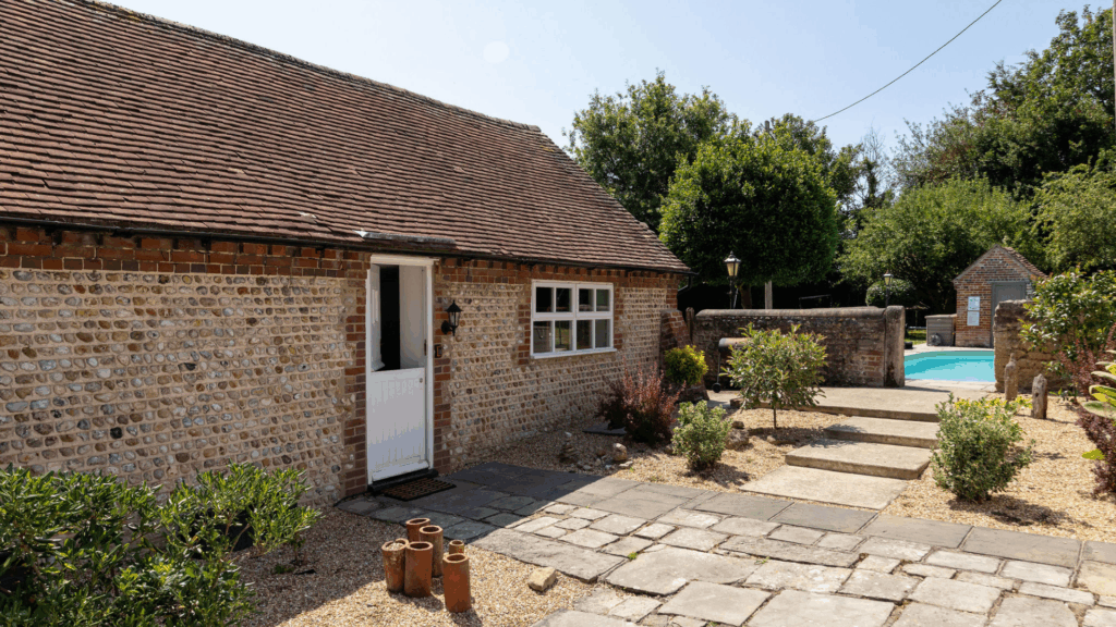 A sun-lit, stone flint cottage annexe with an outdoor swimming pool in the background.
