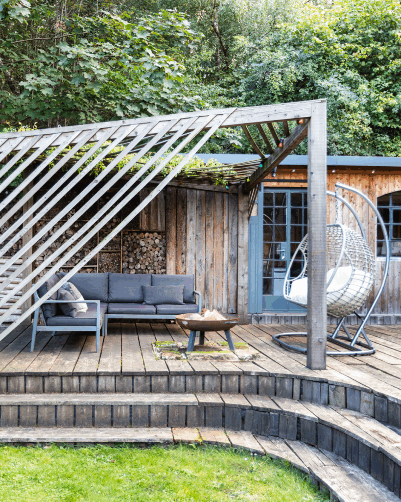 A wooden decking in a garden with outdoor seating and a firepit covered with a pergola.