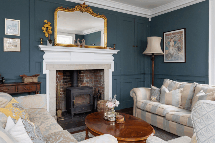 Elegant living room with teal panelled walls, a wood-burning stove in a white fireplace, gold-framed mirror above, striped sofas with cushions, and a wooden coffee table with flowers.