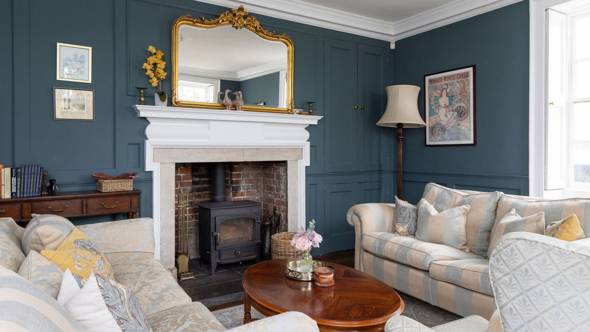 Elegant living room with teal panelled walls, a wood-burning stove in a white fireplace, gold-framed mirror above, striped sofas with cushions, and a wooden coffee table with flowers.