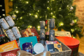 Festive Christmas hamper filled with treats and crackers in front of a decorated Christmas tree with warm lights.