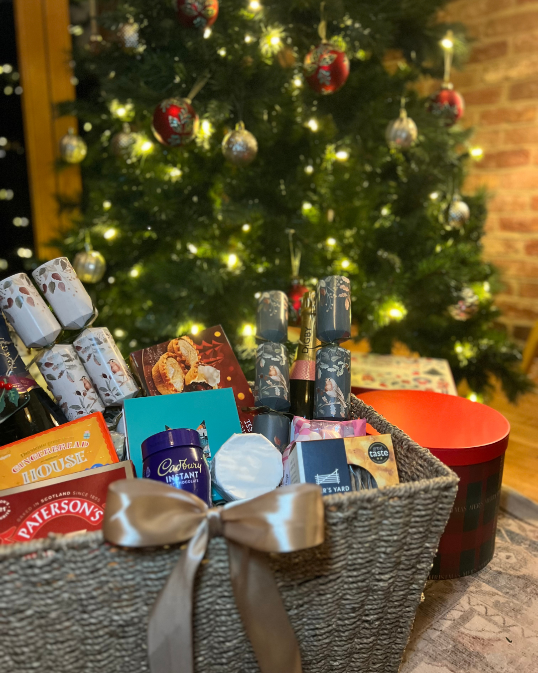 Festive Christmas hamper filled with treats and crackers in front of a decorated Christmas tree with warm lights.