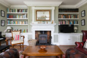 A cosy, elegant living room with built-in bookshelves, a central fireplace, leather armchairs, and a wooden coffee table.