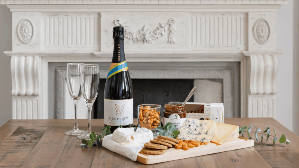 Bottle of Ridgeview sparkling wine with two flutes, cheeses, crackers and chutney arranged on a wooden table in front of a decorative fireplace. Personalised Luxury Staycations.
