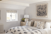 Bright, neutral bedroom with a double bed dressed in blue and white patterned cushions and quilt, bedside lamp, and framed artwork on pale grey walls.