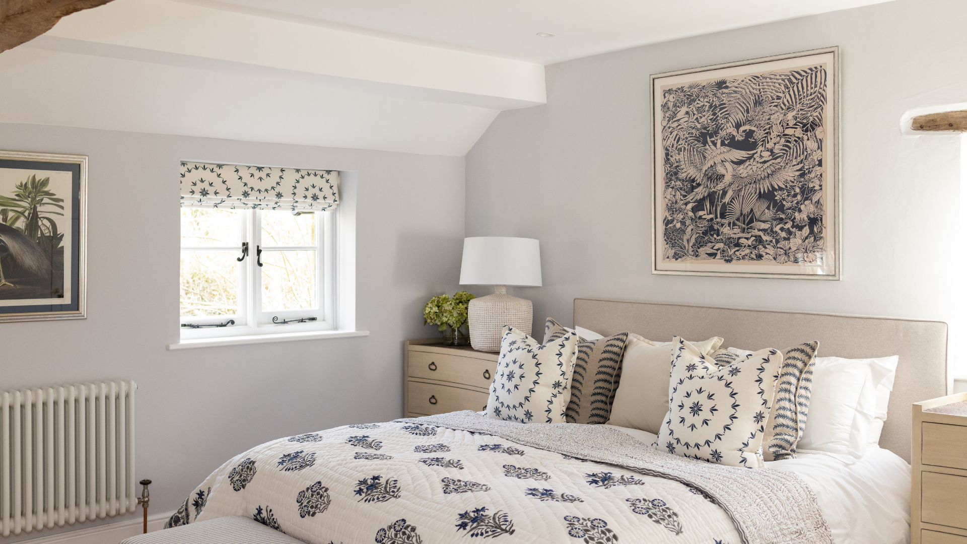 Bright, neutral bedroom with a double bed dressed in blue and white patterned cushions and quilt, bedside lamp, and framed artwork on pale grey walls.
