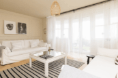 Bright, minimalist living room with neutral tones, white sofas, striped rug and sheer curtains letting in soft natural light.
