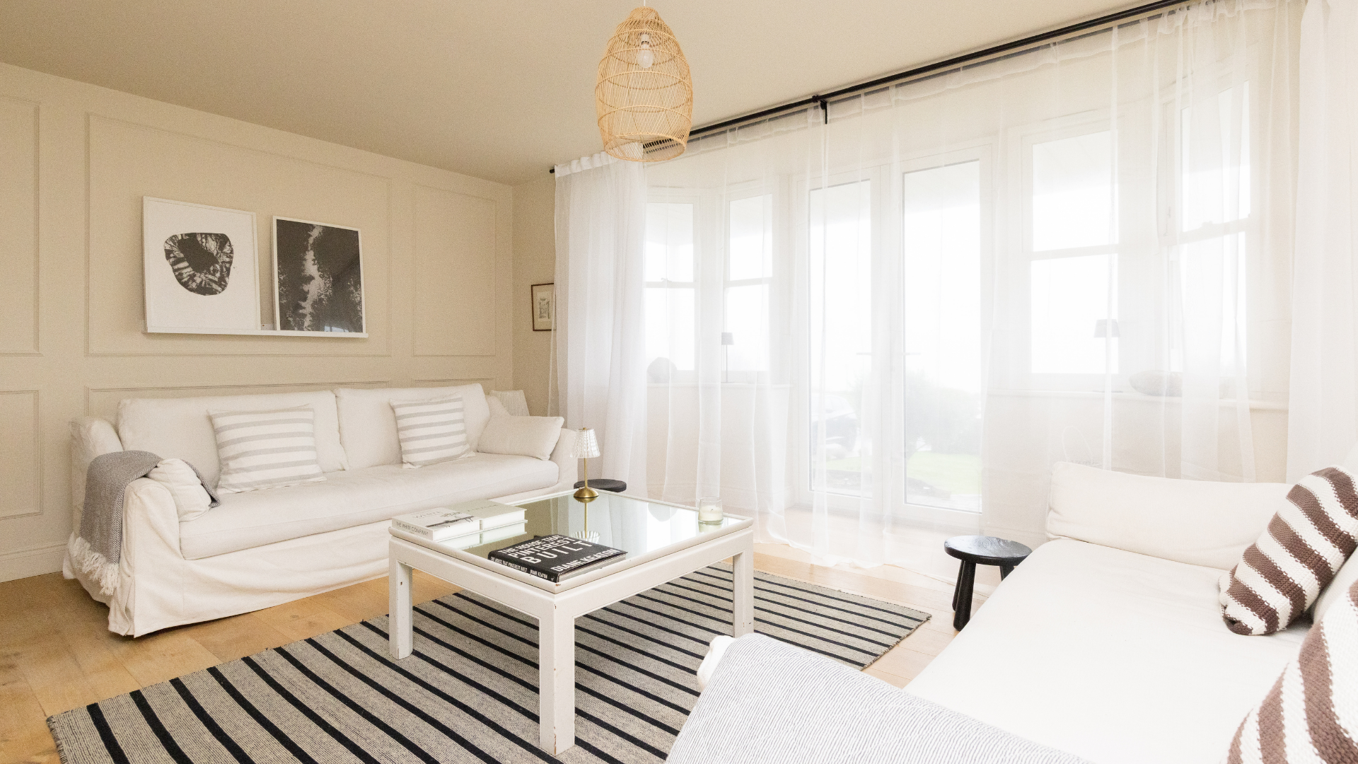 Bright, minimalist living room with neutral tones, white sofas, striped rug and sheer curtains letting in soft natural light.