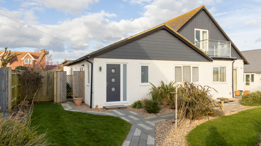 Modern white coastal holiday home with dark cladding, landscaped garden, and balcony under a bright cloudy sky.