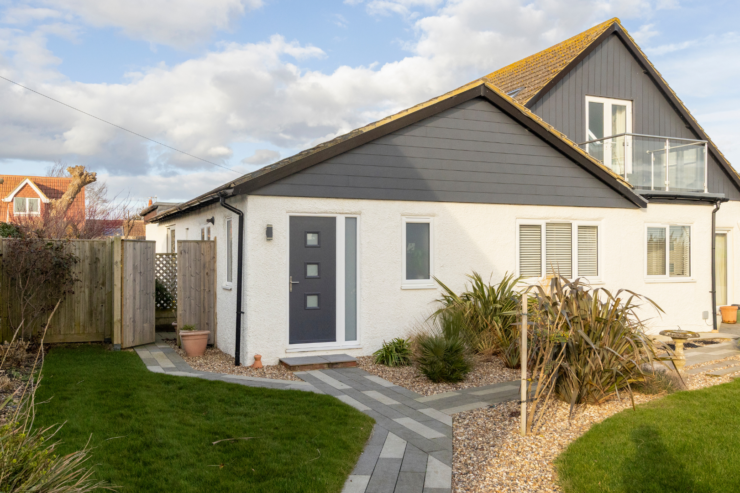 Modern white coastal holiday home with dark cladding, landscaped garden, and balcony under a bright cloudy sky.
