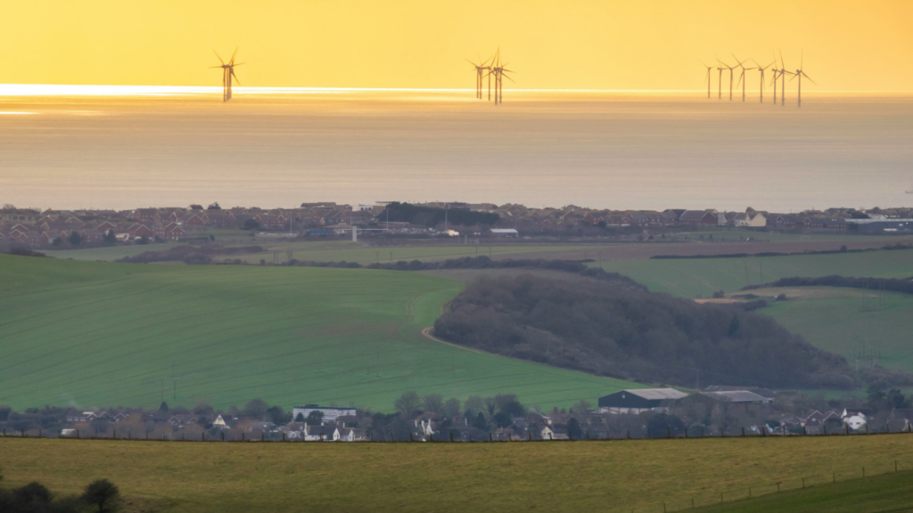 Rolling countryside overlooking a coastal town and offshore wind turbines glowing in golden evening light.