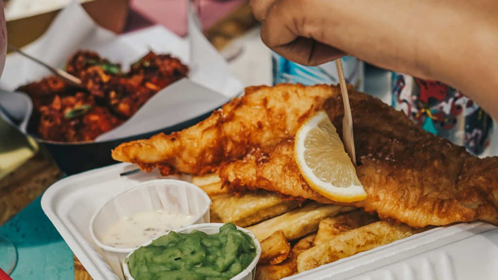 Traditional fish and chips served with mushy peas, tartare sauce, and a lemon wedge on a takeaway tray.