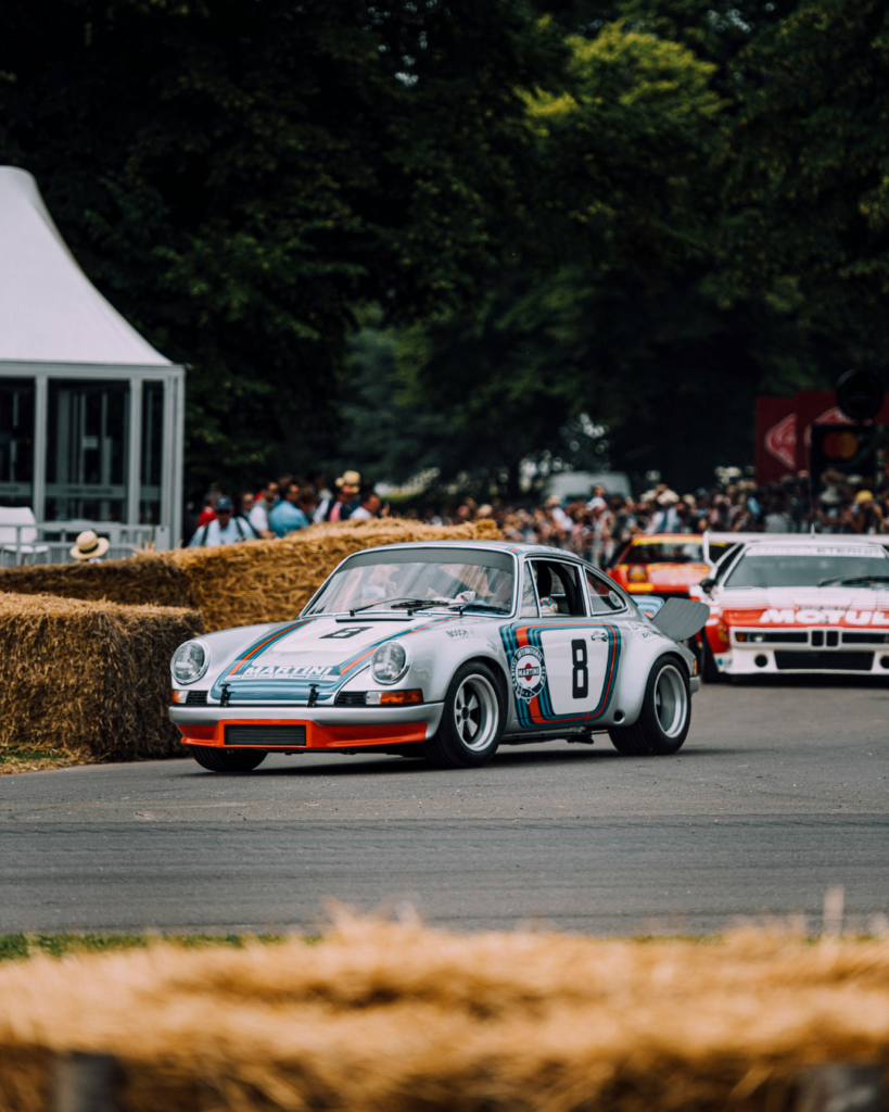 Vintage racing cars navigating a track lined with hay bales during a classic motorsport event, with spectators watching in the background.