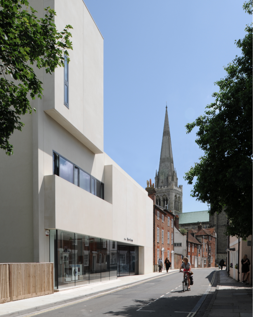 Modern gallery building on a quiet Chichester street, with Chichester Cathedral spire rising in the background on a sunny day.