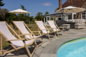 Luxury countryside holiday home with an outdoor swimming pool, striped deck chairs, parasols and patio seating on a sunny day.