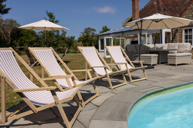 Luxury countryside holiday home with an outdoor swimming pool, striped deck chairs, parasols and patio seating on a sunny day.