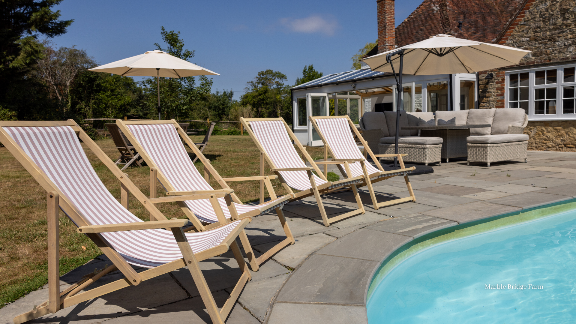 Luxury countryside holiday home with an outdoor swimming pool, striped deck chairs, parasols and patio seating on a sunny day.