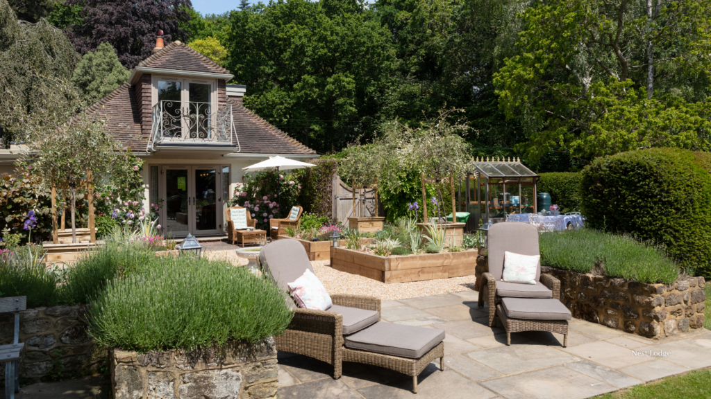 Luxury garden at a countryside holiday cottage with sun loungers, flowering borders, raised planters and outdoor seating surrounded by mature trees.