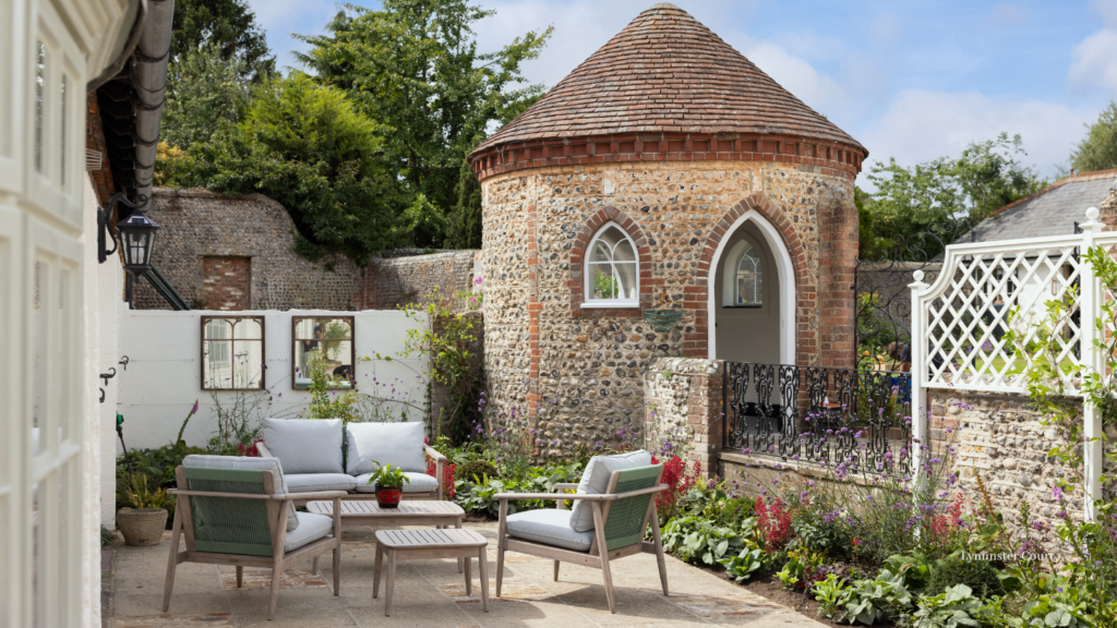 Elegant courtyard garden with outdoor lounge seating beside a historic stone roundhouse, surrounded by colourful flower borders.
